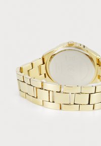 Montre en métal doré avec un bracelet à maillons ; elle présente un boîtier circulaire et une surface en verre lisse au dos.