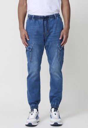 Mężczyzna noszący niebieskie dżinsowe spodnie jogger typu cargo z elastycznym paskiem i ściągaczami, w połączeniu z białymi i niebieskimi sneakersami.