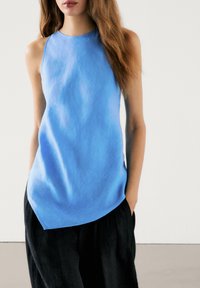 Light blue sleeveless top in a smooth fabric, featuring a high neckline and asymmetric hem, paired with loose black trousers.