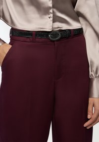 Bordowe dopasowane spodnie w zestawieniu z jasną satynową bluzką. Czarni paski o teksturowanej powierzchni z owalną klamrą dodają detali.