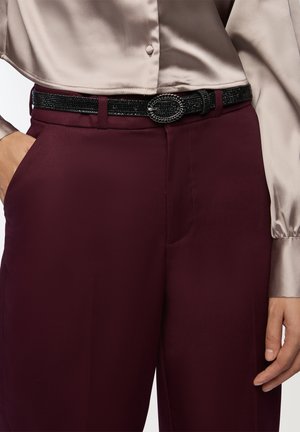 Pantaloni bordeaux su misura abbinati a una blusa di raso leggera. Una cintura nera con superficie texture e fibbia ovale aggiunge un tocco di dettaglio.