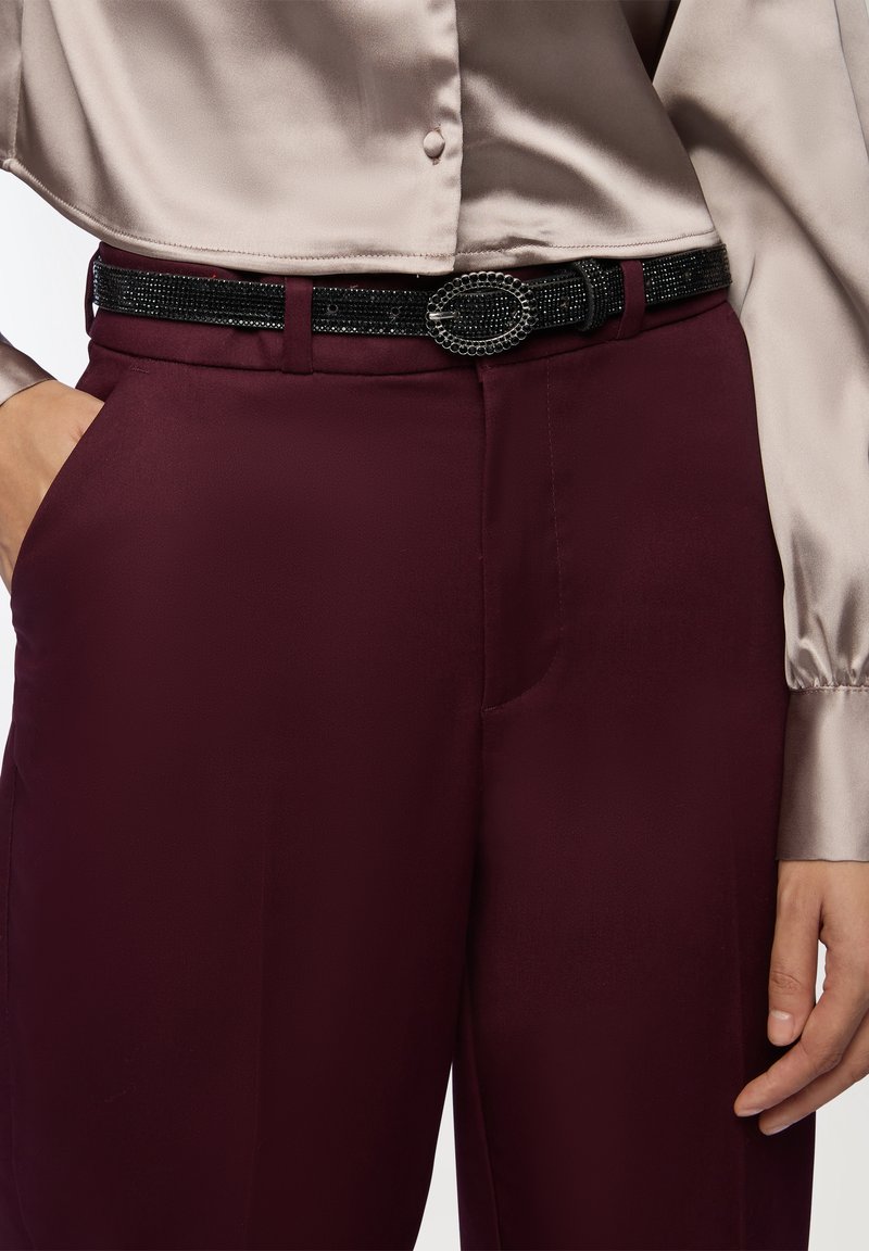 Bordowe dopasowane spodnie w zestawieniu z jasną satynową bluzką. Czarni paski o teksturowanej powierzchni z owalną klamrą dodają detali.