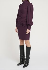 Přiléhavé fialové šaty s dlouhými nabíranými rukávy, shromážděnými detaily na živůtku a délkou po kolena, doplněné černými koženými kozačkami po kolena.