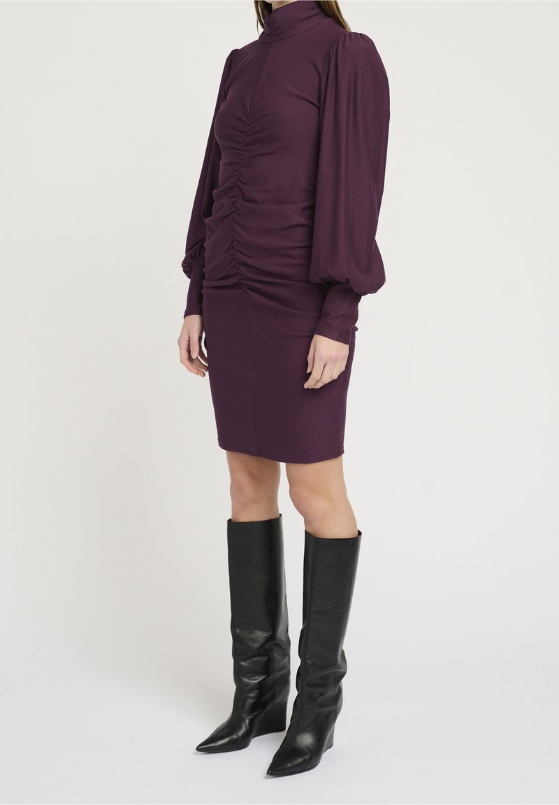 Přiléhavé fialové šaty s dlouhými nabíranými rukávy, shromážděnými detaily na živůtku a délkou po kolena, doplněné černými koženými kozačkami po kolena.
