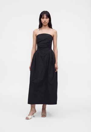 Femme portant une robe midi noire bustier et des sandales blanches à talons, debout devant un fond blanc uni.