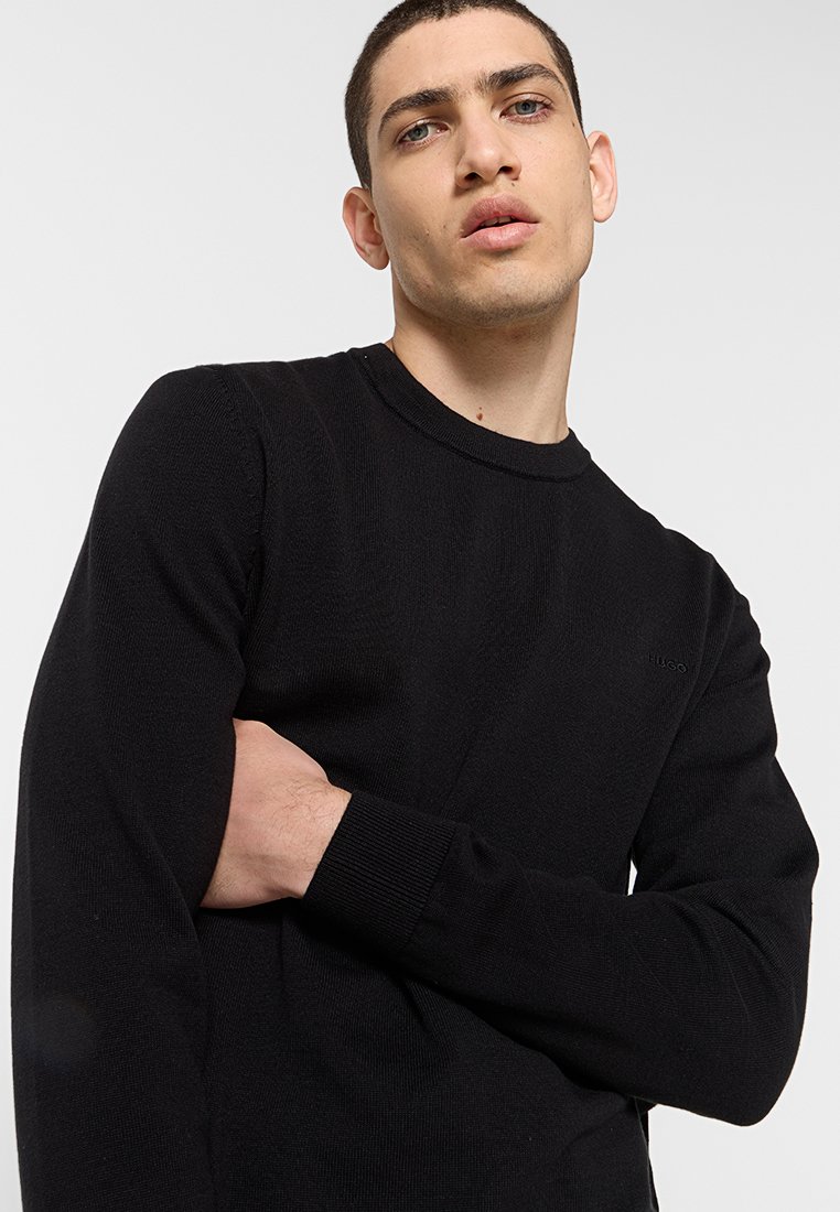 Schwarzer Strickpullover mit Rundhalsausschnitt, gerippten Bündchen und einem dezente Logo. Weicher Stoff und figurbetontes Design.