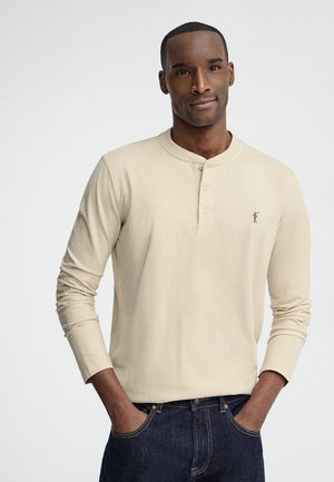 Mężczyzna z rękami w kieszeniach, ubrany w beżową koszulkę henley z długim rękawem i ciemnoniebieskie dżinsy, stojący na jednolitym jasnym tle.