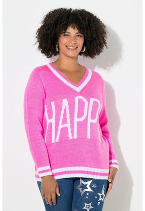 Smilende kvinde med krøllet hår iført en pink sweater med teksten "HAPPY" og jeans dekoreret med sølvstjerne-patches, stående mod en hvid væg.