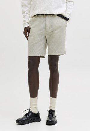 Άνδρας που φοράει μπεζ λινό σορτς, λευκό πουκάμισο με μακριά μανίκια, μαύρη πλεκτή ζώνη, κρεμ κάλτσες και μαύρα δερμάτινα δερμάτινα παπούτσια με κορδόνια.