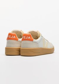 Zapatillas deportivas blancas y beige con acentos naranjas en el talón, materiales de ante y cuero, suela de goma elevada y marca "VEJA" en el talón.