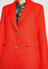 Blazer rouge vif texturé avec de larges revers, deux boutons dorés et des poches à rabat, porté sur un chemisier noir et gris à motifs.
