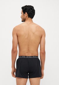 Des boxers en coton noir présentent une ceinture élastique avec logo. Le mannequin est vu de dos, mettant en valeur la coupe et la longueur des shorts.
