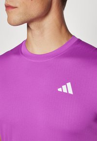 Camiseta atlética morada con cuello redondo y mangas cortas, que presenta una tela texturizada y un logotipo blanco en la parte izquierda del pecho.