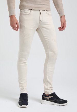 Uomo che indossa jeans slim-fit color avorio slavato e sneakers nere e beige, in piedi davanti a uno sfondo semplice.