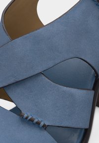 Μπλε σουέντ σανδάλι με σταυρωτούς ιμάντες και λεπτομέρεια ορατών ραφών σε ανοιχτό φόντο.