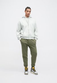 Grauer Reißverschluss-Hoodie mit Fronttaschen, kombiniert mit olivgrünen Jogginghosen. Trägt dazu schwarze und weiße High-Top-Sneaker mit gelben Akzenten.