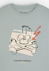 Camiseta de algodón azul claro con un gráfico de calavera y tibias cruzadas en negro con acentos rojos, y salpicaduras de pintura blanca debajo del diseño.