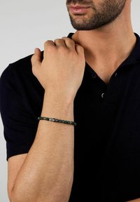 Bracciale con perline nere, pietre verde scuro e un ciondolo centrale nero con un motivo bianco, indossato su un polso nudo.