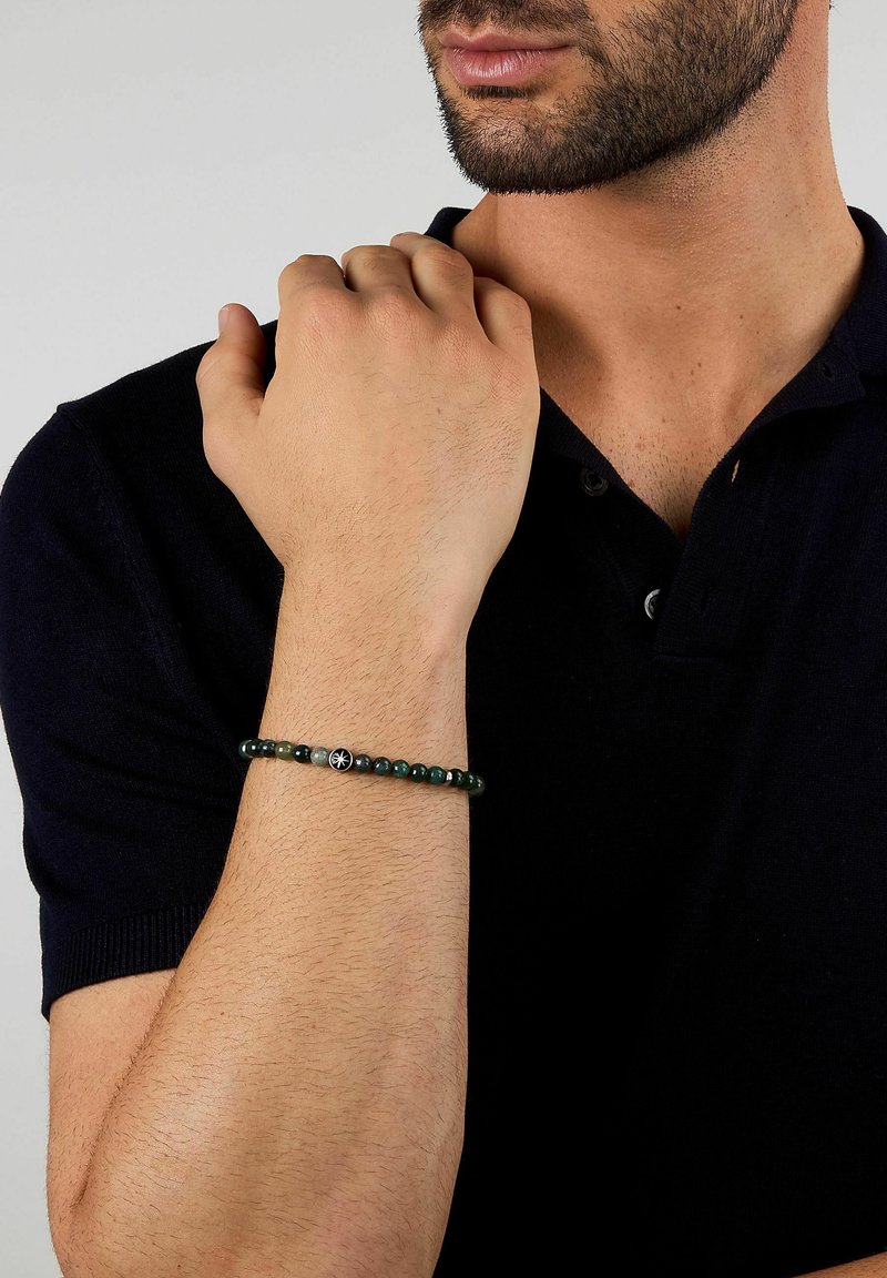 Bracciale con perline nere, pietre verde scuro e un ciondolo centrale nero con un motivo bianco, indossato su un polso nudo.