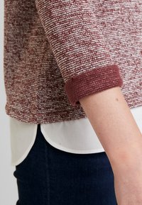 Suéter de punto burdeos texturizado con puños enrollados, superpuesto a una camisa blanca. Unos jeans oscuros ajustados completan el atuendo.