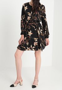 Donna che indossa un vestito lungo a maniche lunghe di colore nero con motivo floreale, con vita stretta e scarpe a blocco beige con punte nere.