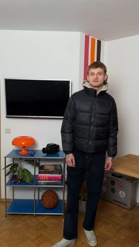 Νέος άνδρας με μαύρο μπουφάν και λευκά αθλητικά παπούτσια στέκεται σε γωνιακό δωμάτιο με τηλεόραση στον τοίχο, πορτοκαλί λάμπα, κάμερα και μπάλα του μπάσκετ σε ράφια.