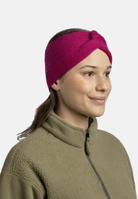 Gestricktes rosa Stirnband mit einem verdrehten Knotendesign. Trägt es über einer hellbraunen Fleecejacke, die die glatte Textur und die lebendige Farbe hervorhebt.