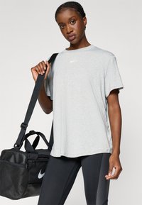 Ανοιχτό γκρι κοντομάνικο μπλουζάκι Nike με λαιμόκοψη. Μαύρη τσάντα αθλητικών ειδών Nike με υφασμάτινες λεπτομέρειες, φορεμένη στον ώμο με μαύρες κολάν.