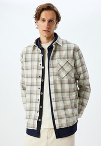 Camisa de franela a cuadros en beige con detalles en marrón y azul marino, que presenta un cierre de botones, cuello con puntas y un bolsillo en el pecho.