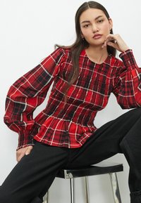 Top à carreaux rouge et noir avec corsage froncé et manches bouffantes. Il présente un col rond et un ourlet peplum froncé. Les accessoires comprennent de petites boucles d'oreilles.