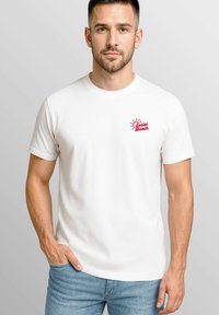 Weißes Baumwoll-T-Shirt mit kurzen Ärmeln, ausgestattet mit einem kleinen roten "Social Beach"-Logo auf der linken Brustseite. Getragen mit hellblauen Jeans.