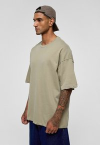 Olijfgroene oversized katoenen T-shirt met een ronde halslijn en verlaagde schouders, gedragen met donkere denim shorts. Heeft zichtbare tatoeages op de arm.