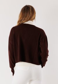 Donna con capelli lunghi che indossa un maglione corto marrone scuro sopra una camicia bianca e pantaloni bianchi, vista da dietro su uno sfondo neutro.