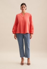 Korale blouse z długimi, haftowanymi rękawami, zapinana na guziki, z dekoltem w kształcie litery V, zestawiona z jasnoniebieskimi prostymi dżinsami i czarnymi sandałami na paski.