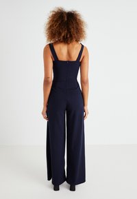 Navy jumpsuit with wide leg, fitted bodice, and thick straps. Features a back zipper and smooth, textured fabric.