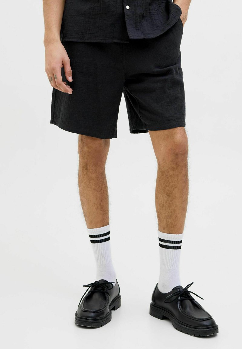 Pantalones cortos de tela negra con un acabado texturizado, combinados con calcetines blancos que presentan dos rayas negras y zapatos negros con cordones y una suela gruesa.