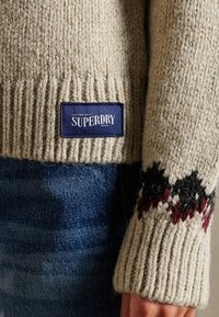 Beige neulepaita, jossa on ribbitetty pinta, ja siinä on tummansininen etiketti, jossa lukee "Superdry". Hihansuiden reunassa on musta- ja viininpunainen sahalaitakuvio.