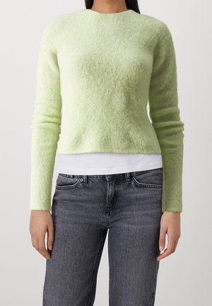 Femme portant un pull duveteux vert clair à manches longues sur une chemise blanche, associée à un jean gris foncé taille haute, debout devant un fond uni.