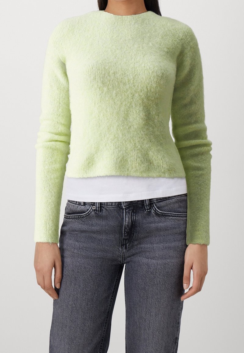 Femme portant un pull duveteux vert clair à manches longues sur une chemise blanche, associée à un jean gris foncé taille haute, debout devant un fond uni.