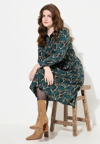 Abito verde scuro con una fantasia di rami d'albero, colletto, maniche lunghe e vita arr gathered. Indossato con stivali alla caviglia beige in suede e collant scuri.