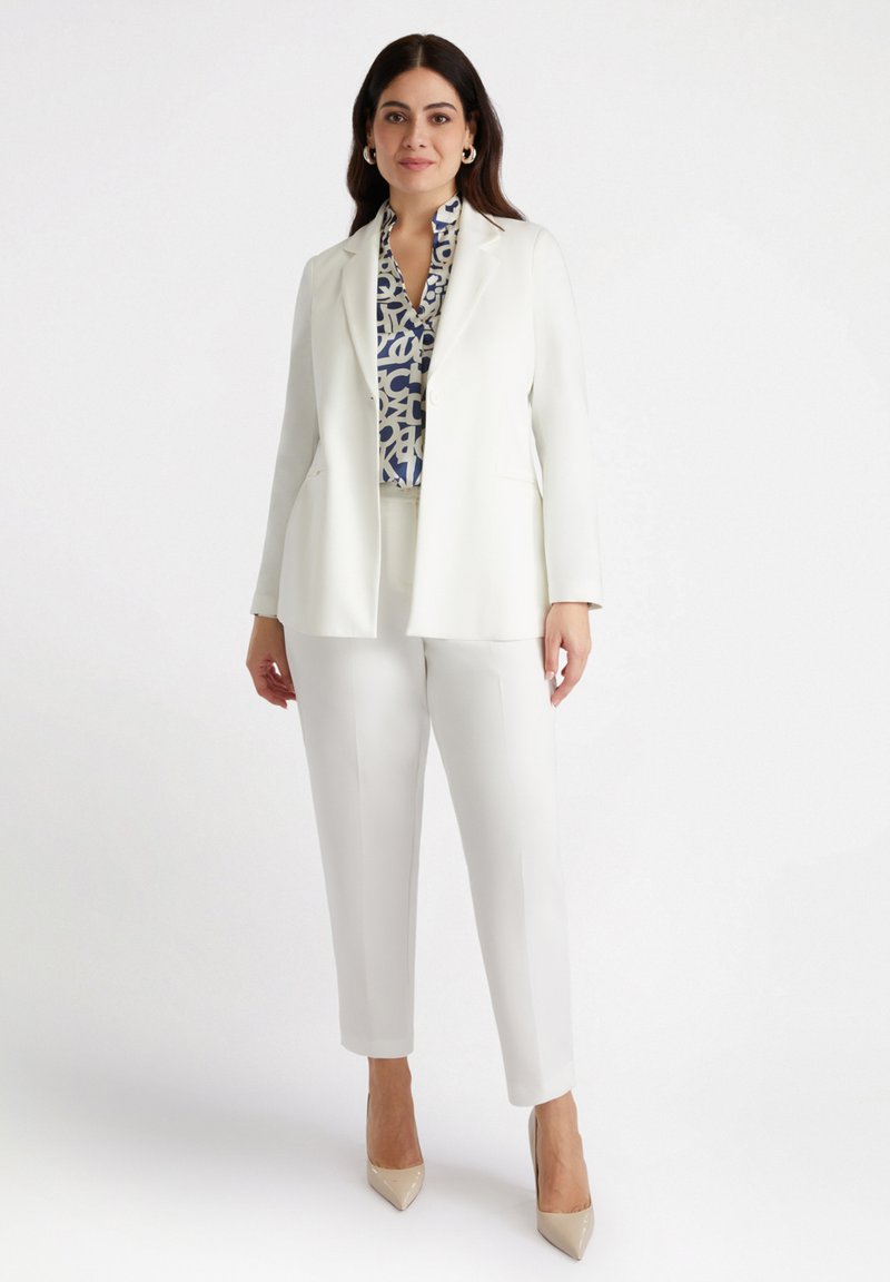 Biała marynarka z klapami, w połączeniu z białymi spodniami i wzorzystą niebieską bluzką. Modelka w beżowych szpilkach.