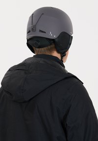 Grå skid hjälm med ventilationsöppningar och justerbart hakband, bärs under en svart huva jacka. Matt yta med diskret branding på baksidan.