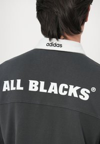 Siva dolgimi rokavi sive barve z belim ovratnikom, na hrbtni strani z boldiranimi belimi črkami "ALL BLACKS" in logotipom "adidas" na ovratniku.