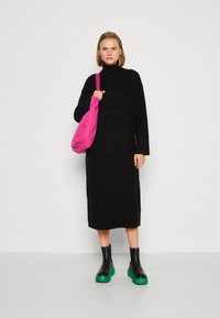 Abito lungo nero con maniche a lunghe e collo alto; modella che tiene una borsa rosa; indossa stivaletti neri con suole a piattaforma verde.