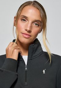 Svart zip-up sweatshirt med hög krage, tillverkad av mjukt material. Har en liten logotyp på bröstet och en kontrasterande dragkedja.