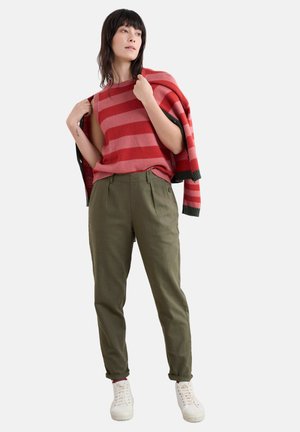 Un pull sans manches rayé rouge et rose assorti à un pantalon vert olive. Le mannequin tient un cardigan rayé rouge et noir. Baskets blanches.