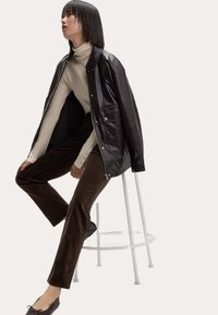 Braune Lederjacke über einem beigen Rollkragenpullover, kombiniert mit braunen Cordhosen. Modell sitzt auf einem weißen Hocker. Sanfte Texturen und neutrale Töne.