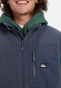 Man som ler och har på sig en grön huvtröja under en marinblå zip-up fleecejacka med bröstficka och logotyp.