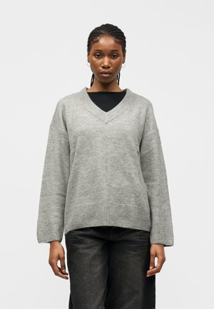 Kvinde med flettet hår iført grå V-hals sweater over sort top og mørke jeans, stående mod en ensfarvet hvid baggrund.