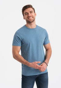 Camiseta azul de manga corta con pequeños lunares oscuros, cuello redondo y aberturas laterales. La tela parece suave y transpirable.